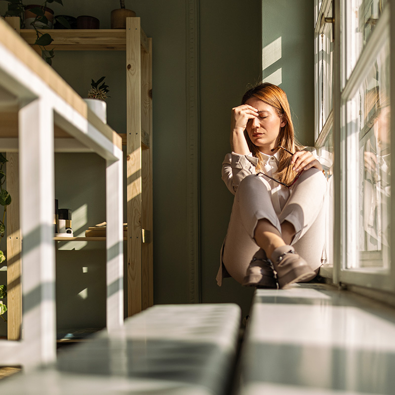 Depressed young businesswoman sitting on window sill holding her hand